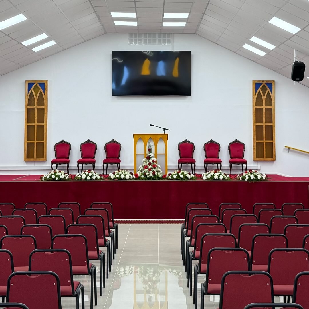Interior del templo: púlpito de madera con arcos, sillas de respaldo rojo en el altar, arreglos de flores blancas y rojas, y filas de asientos frente al escenario